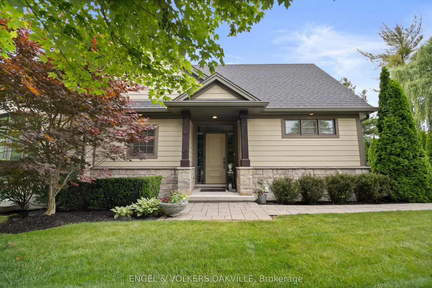 Front view of a beige house with a gray roof, stone accents, and a well-manicured lawn. A paved walkway leads to the front door.