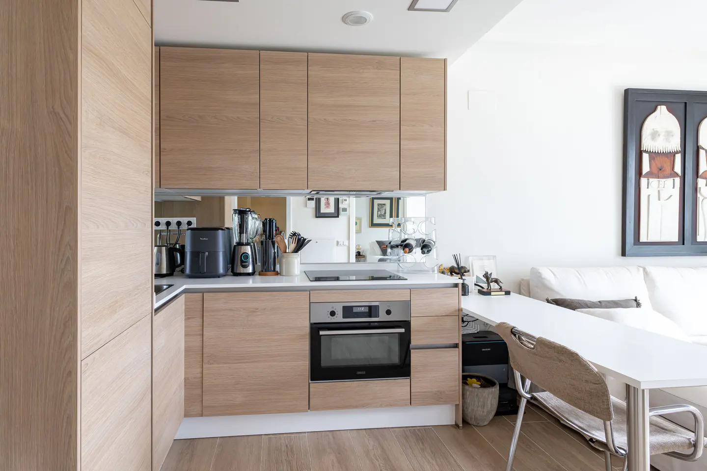 Modern kitchen with light wood cabinets, white countertops, and stainless steel appliances. A white table and chair are visible in the foreground.