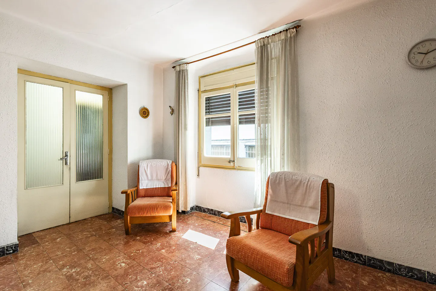 A bright room with two orange armchairs, a window with sheer curtains, and a glass-paneled door. The floor is red tile.
