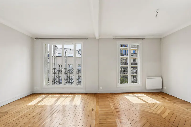 Bright, empty room with white walls, herringbone wood floor, and two windows showing city buildings.