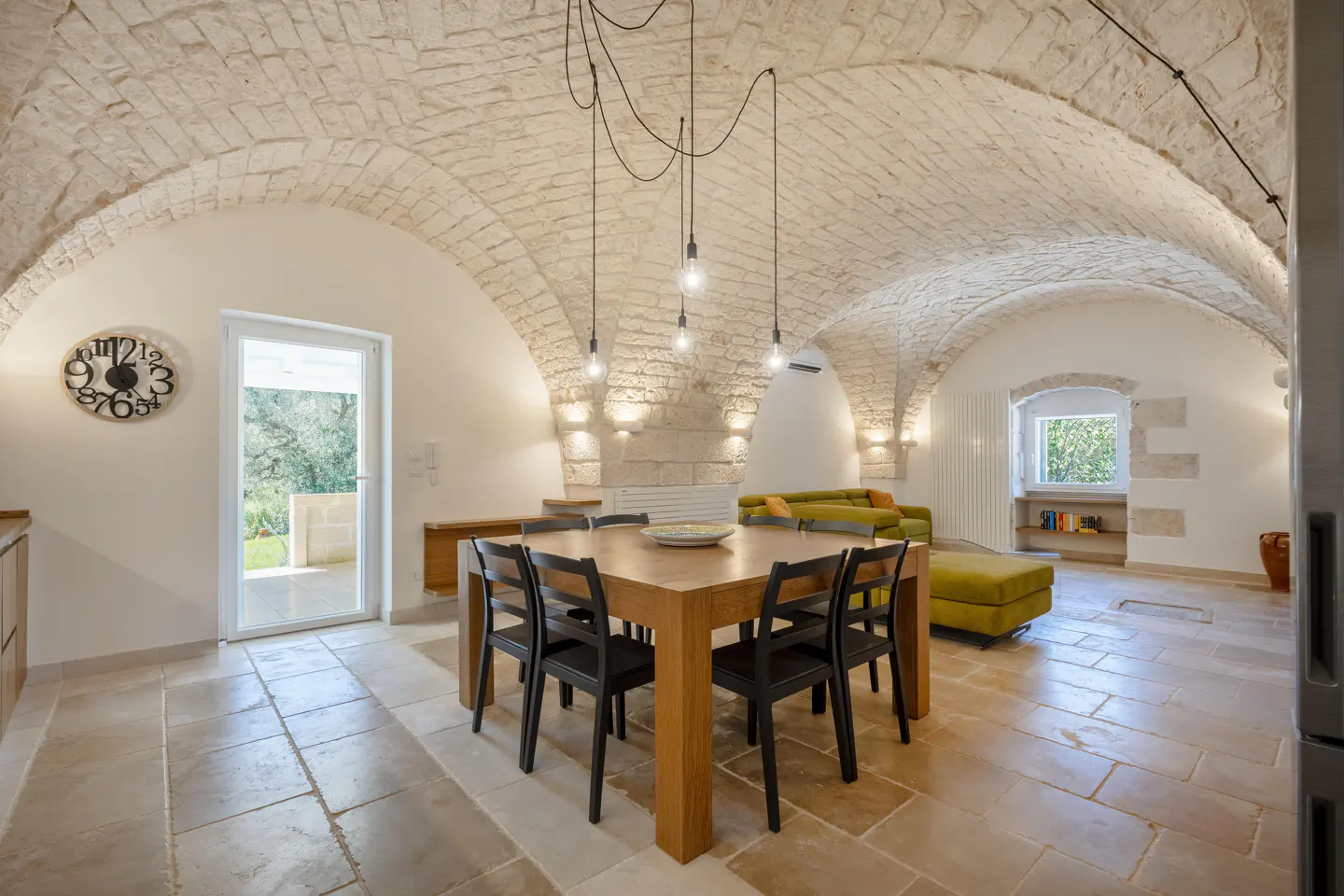 Interior view of a dining room with stone floors, a vaulted ceiling, and a wooden table with black chairs. A green sofa is in the background.