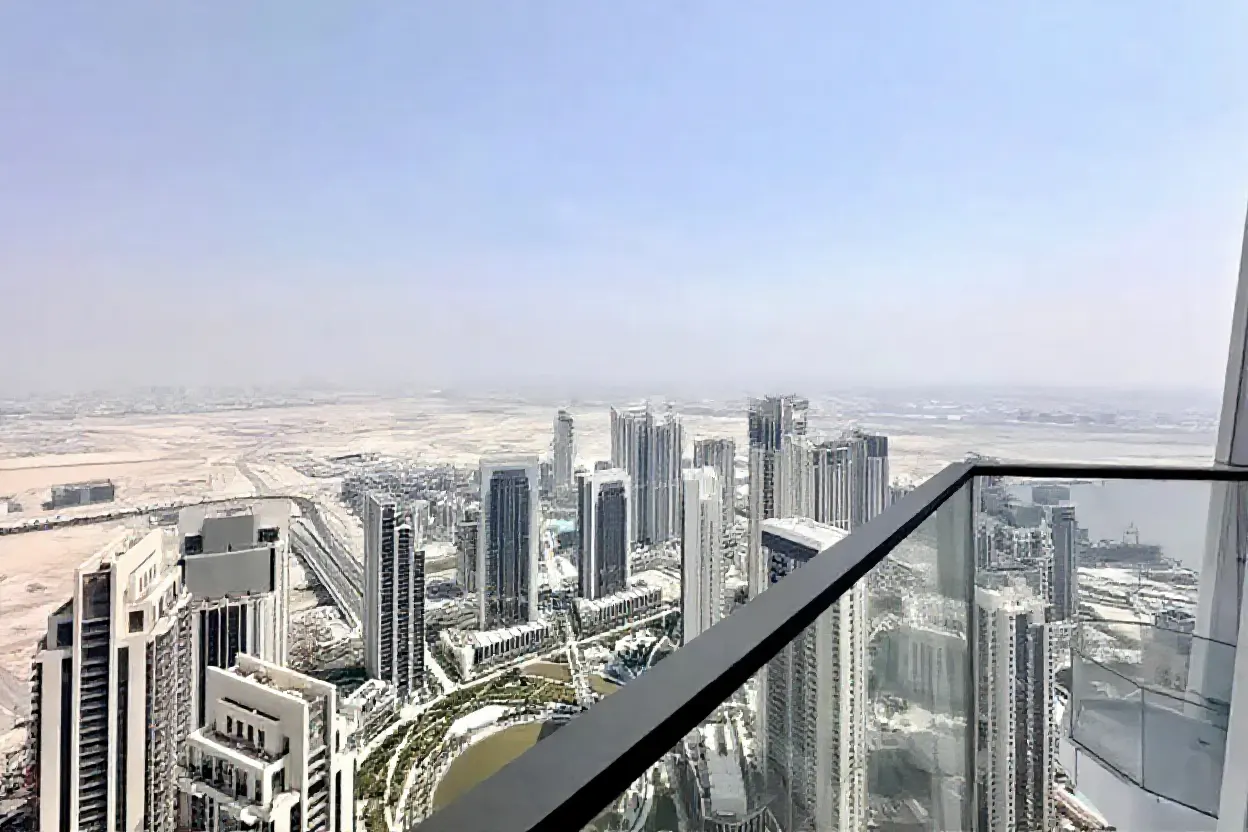 View from a high-rise balcony overlooking a city with modern buildings and a desert landscape under a hazy blue sky.