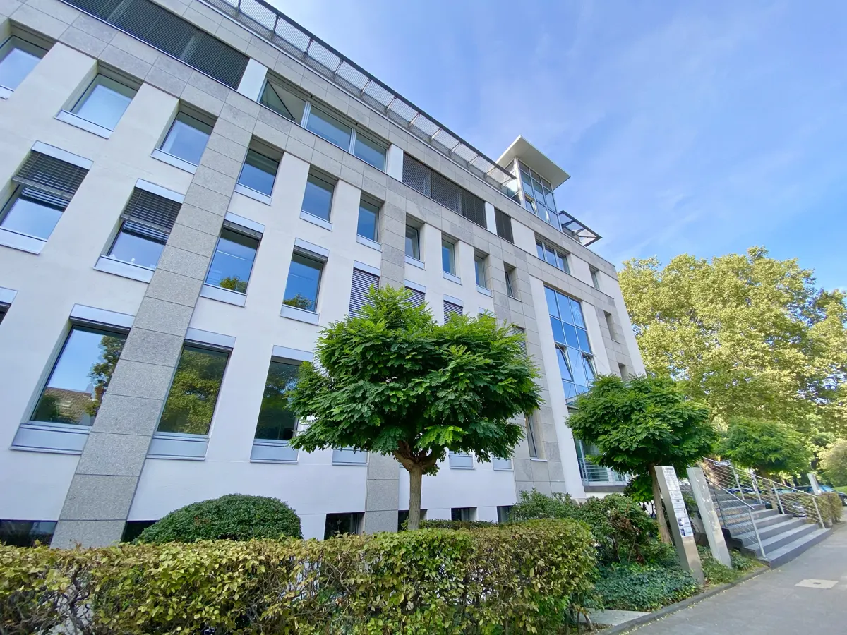 Exterior view of a modern, multi-story office building with white facade, gray accents, and many windows, framed by green trees and bushes.