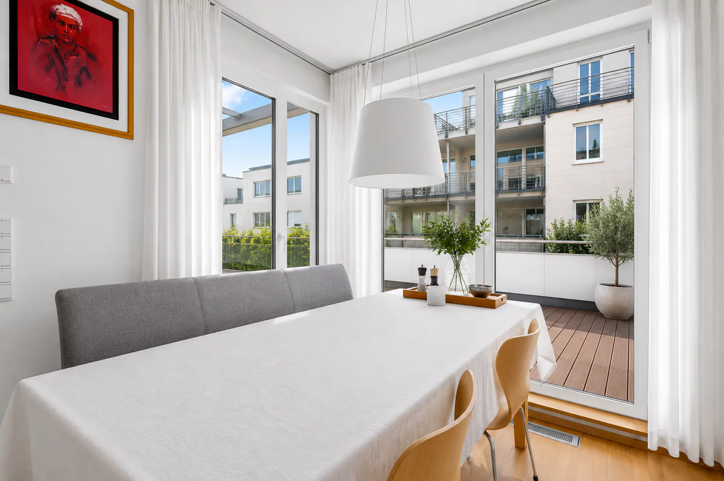 Dining area with a white-clothed table, grey bench, wooden chairs, and a balcony view through large glass doors.