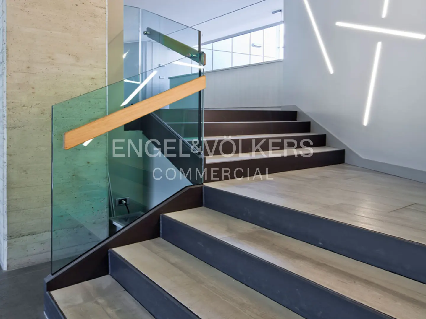 Modern staircase with light wood treads, dark risers, and a glass railing with a wooden handrail in a bright office lobby.