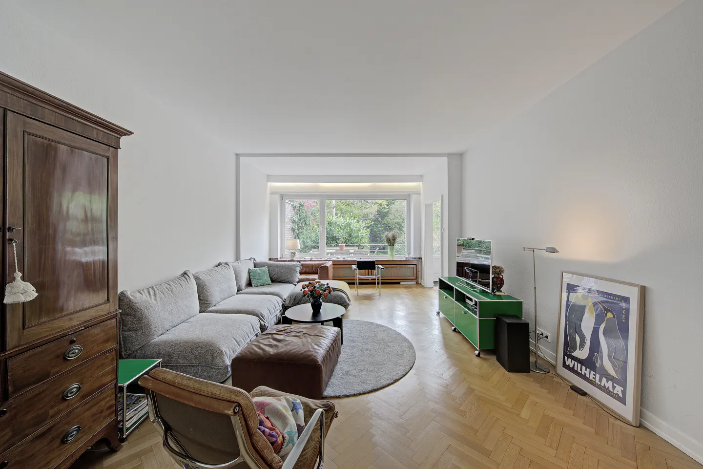 Bright living room with herringbone wood floors, a gray sectional sofa, and a large window overlooking greenery. A green cabinet and penguin art add pops of color.