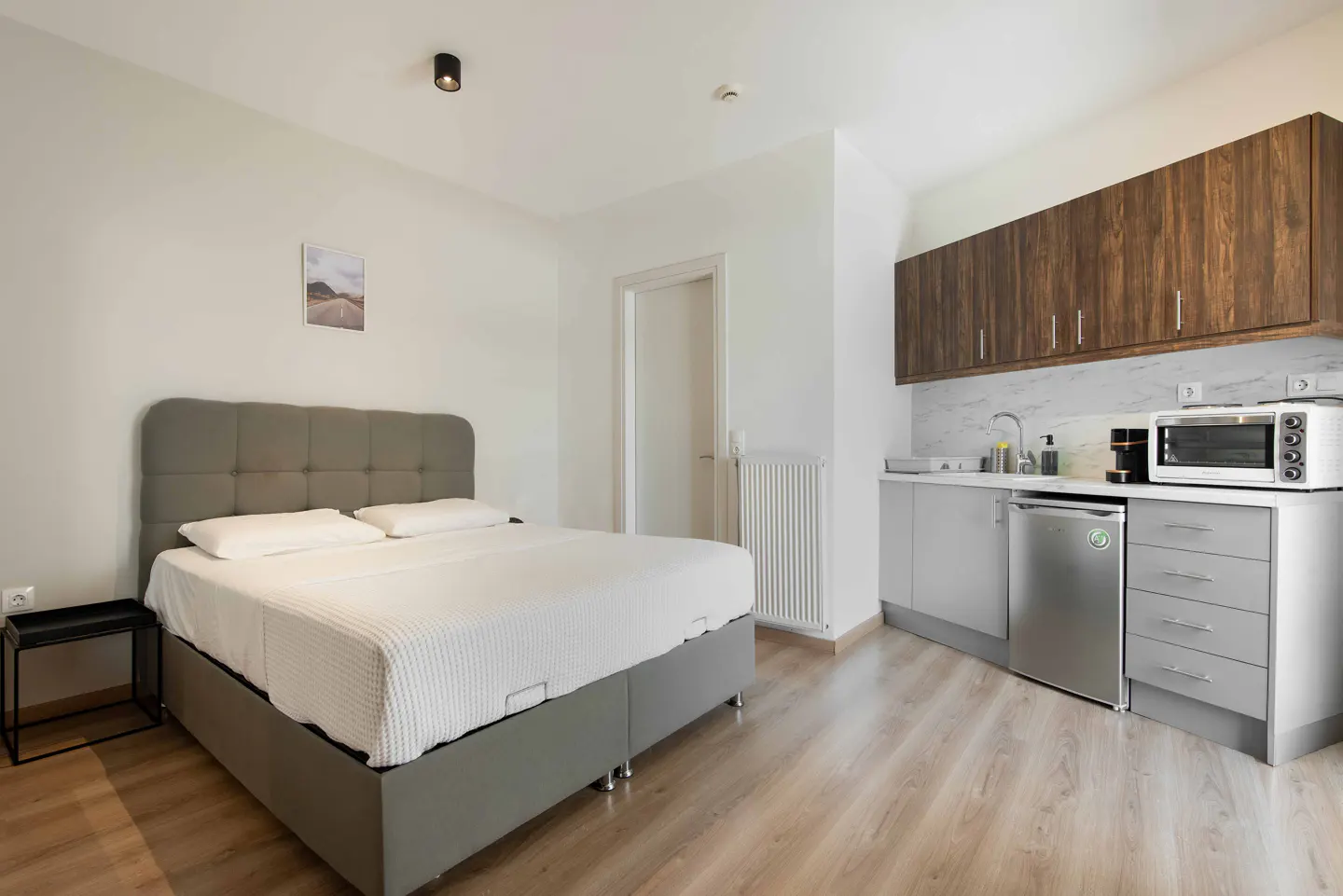 Studio apartment with a gray bed, white linens, and a kitchenette with wood cabinets and gray counters.