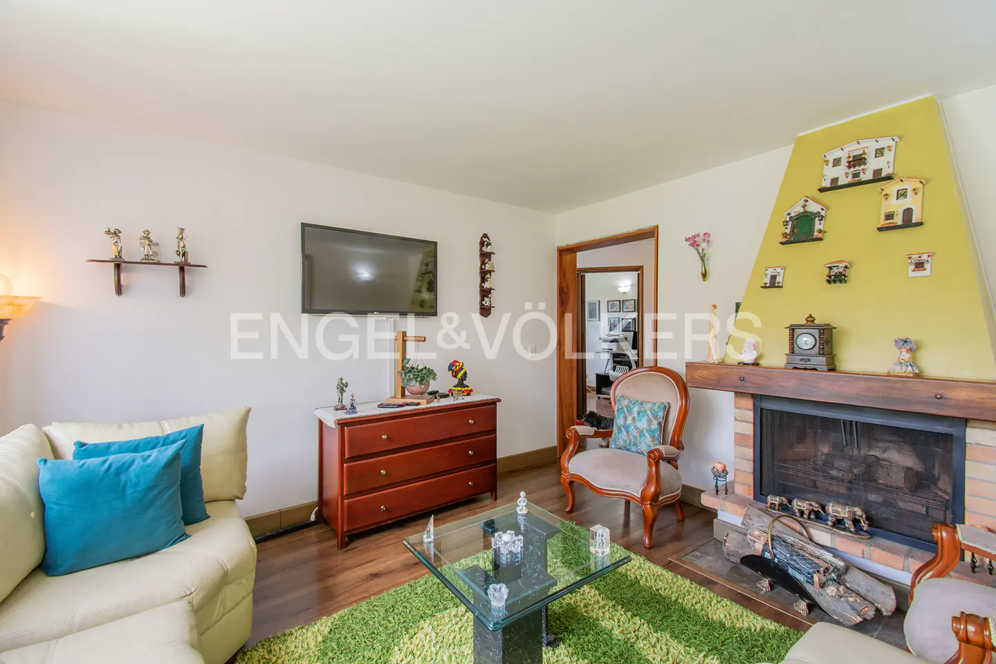 Living room with a fireplace, a cream sofa with blue pillows, a wooden dresser, and a glass coffee table on a green rug.