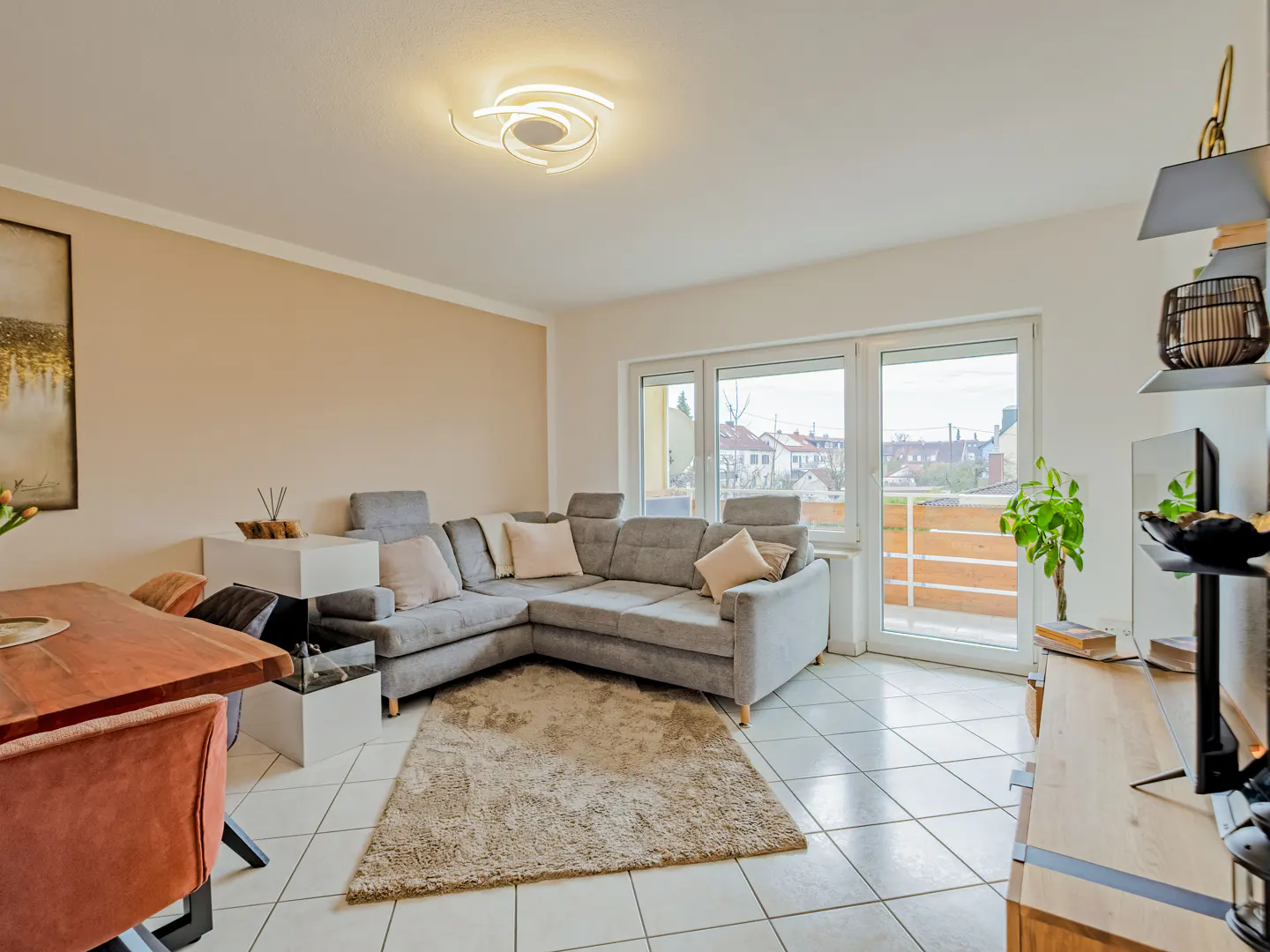 Bright living room with a gray sectional sofa, beige rug, and white tile floor. Large windows overlook a balcony. A wooden table and chairs are visible.