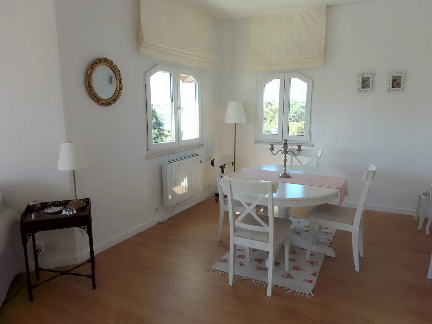 Bright dining room with white walls, wood floors, and white furniture. Windows with cream-colored shades let in natural light.