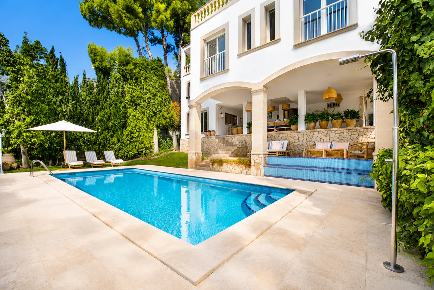 A bright blue swimming pool sits in front of a white villa with a covered patio, lounge chairs, and lush green trees.