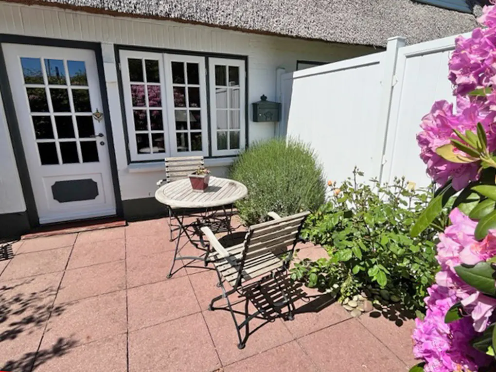 A patio with a small round table and two chairs, set against a white house with a thatched roof and pink flowers.