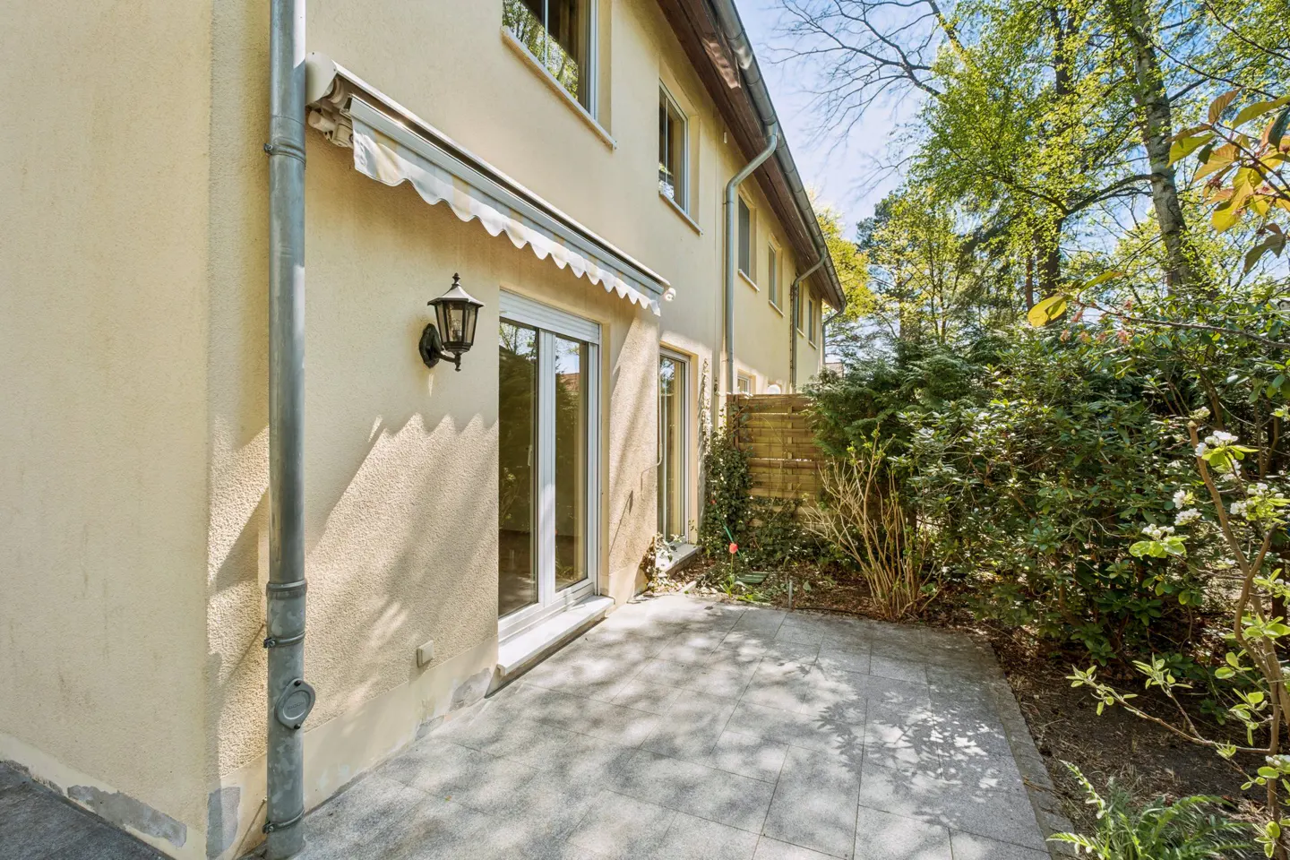 Exterior view of a two-story beige house with a stone patio, sliding glass doors, and green foliage.