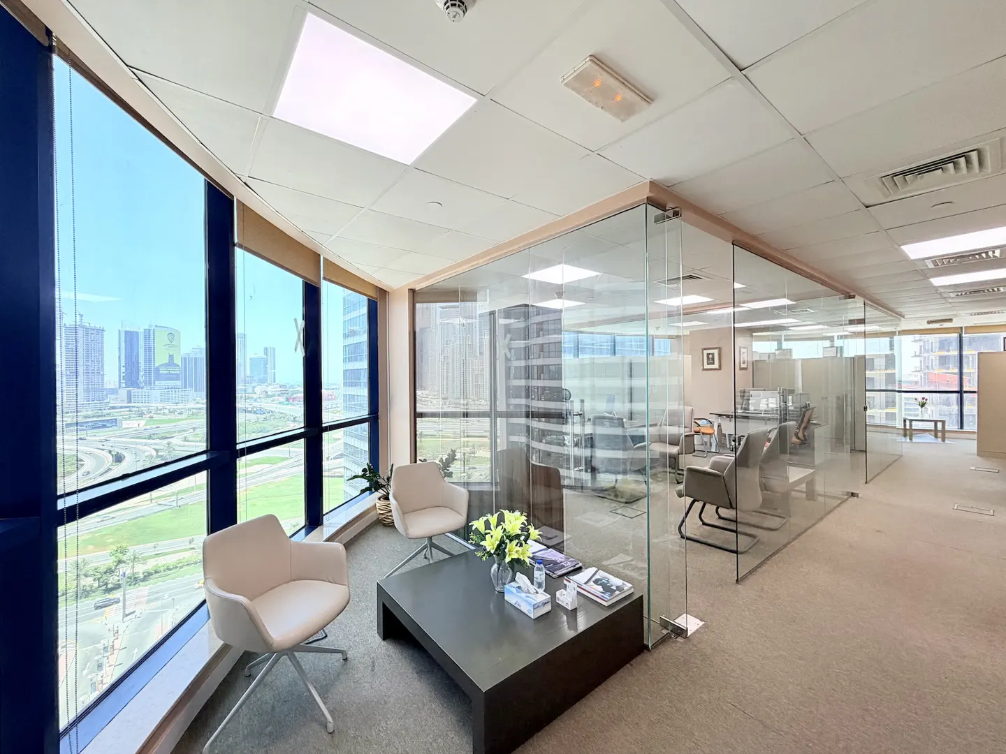 Office interior with two white chairs, a dark coffee table, and a glass-walled conference room, with a city view through large windows.