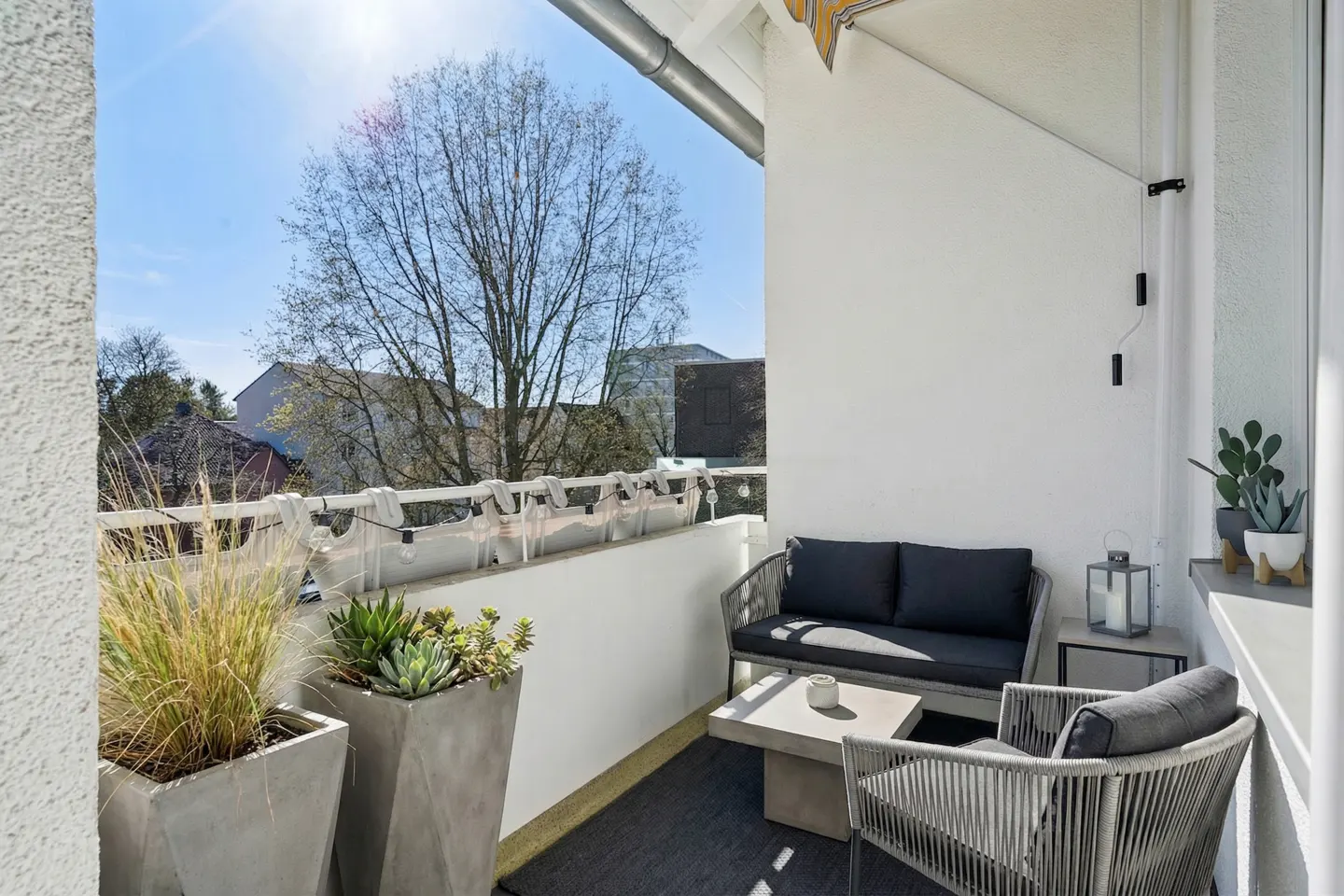 A sunlit balcony with gray woven furniture, including a loveseat and chair, on a dark rug. Potted plants line the white railing.