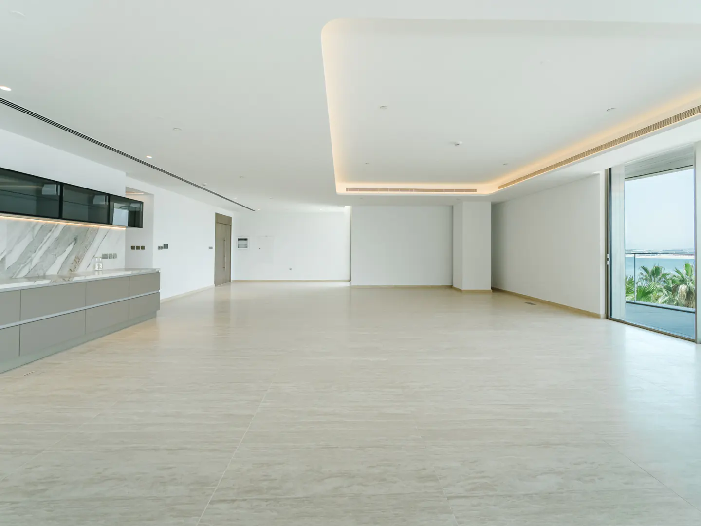 Bright, empty room with beige tile floor, white walls, and recessed lighting. A gray kitchen counter is on the left, and a sliding glass door shows palm trees and ocean.