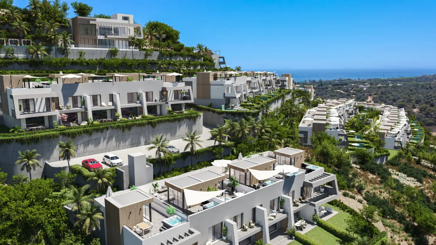 Aerial view of modern white hillside villas with palm trees, green foliage, and ocean view under a clear blue sky.
