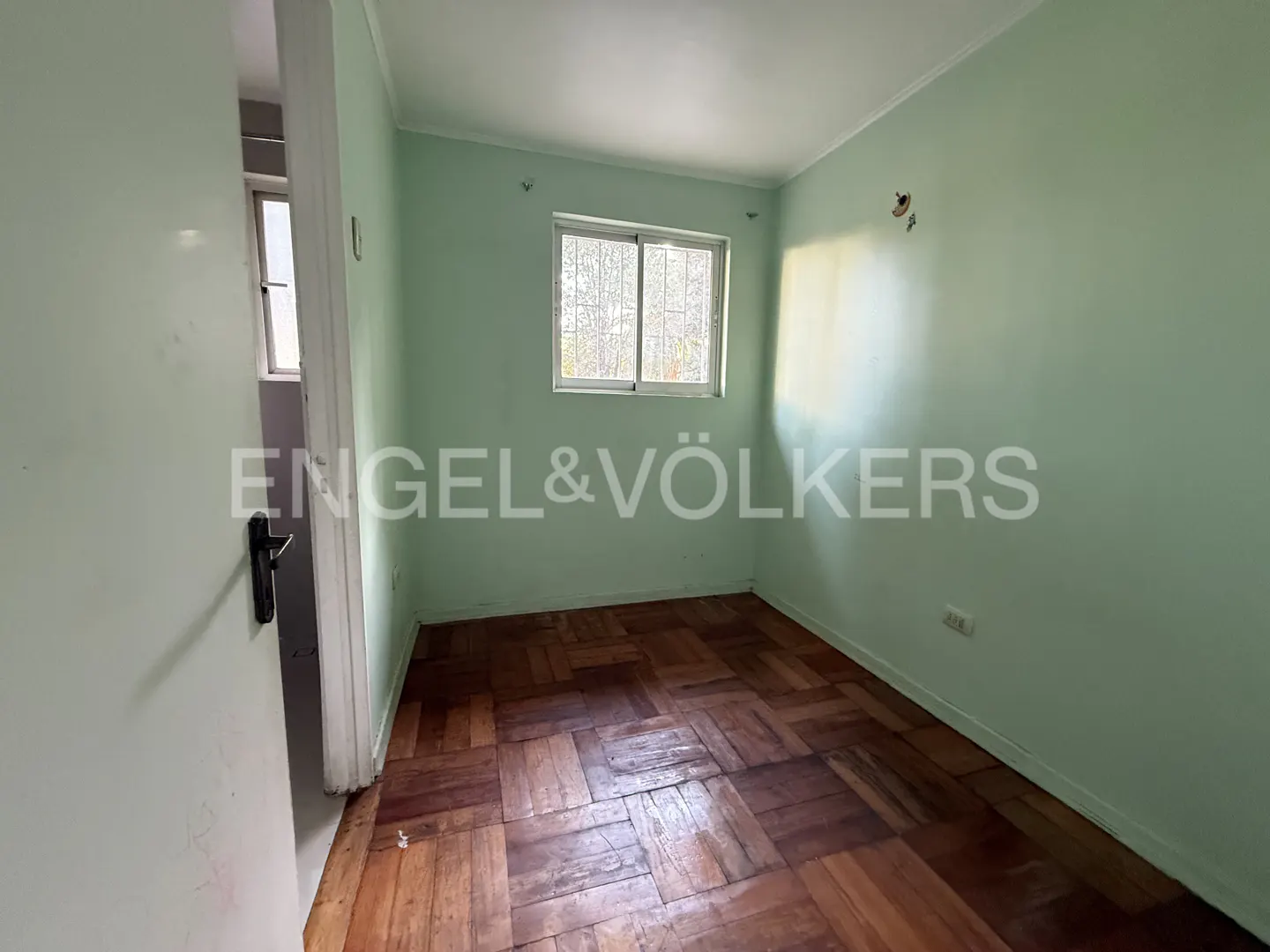 Empty room with light green walls, wood floors, and a window. Doorway on the left. Engel & Völkers logo visible.