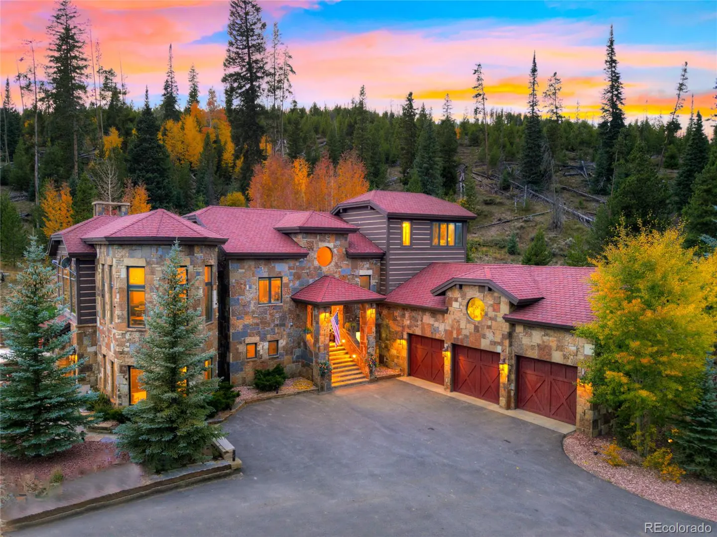 Stone mansion with red roof and three-car garage, set against a forest and colorful sunset sky.