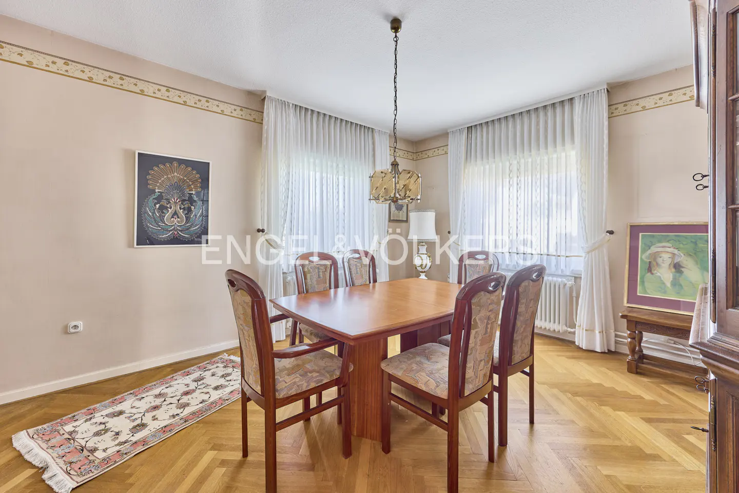 Dining room with a wooden table, four patterned chairs, herringbone floors, sheer curtains, and framed wall art.