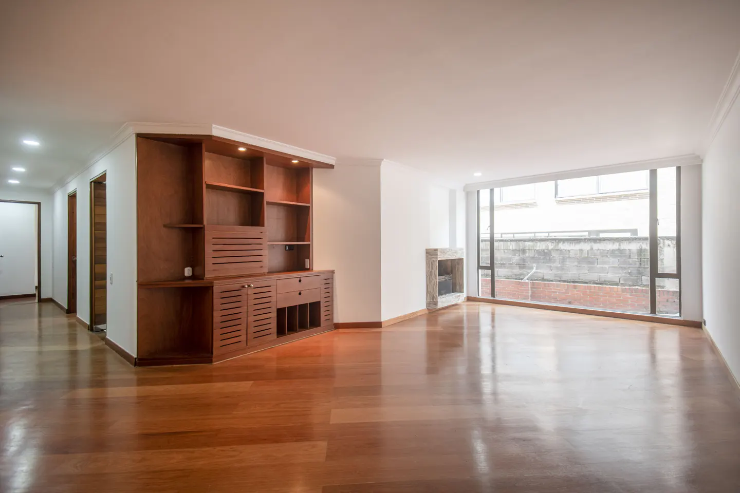 Bright, empty living room with hardwood floors, a built-in wooden cabinet, and a stone fireplace near a large window.