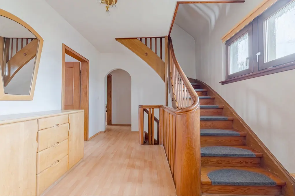 Bright hallway with wood floors, a curved wood staircase with gray treads, and a light wood dresser with a mirror.