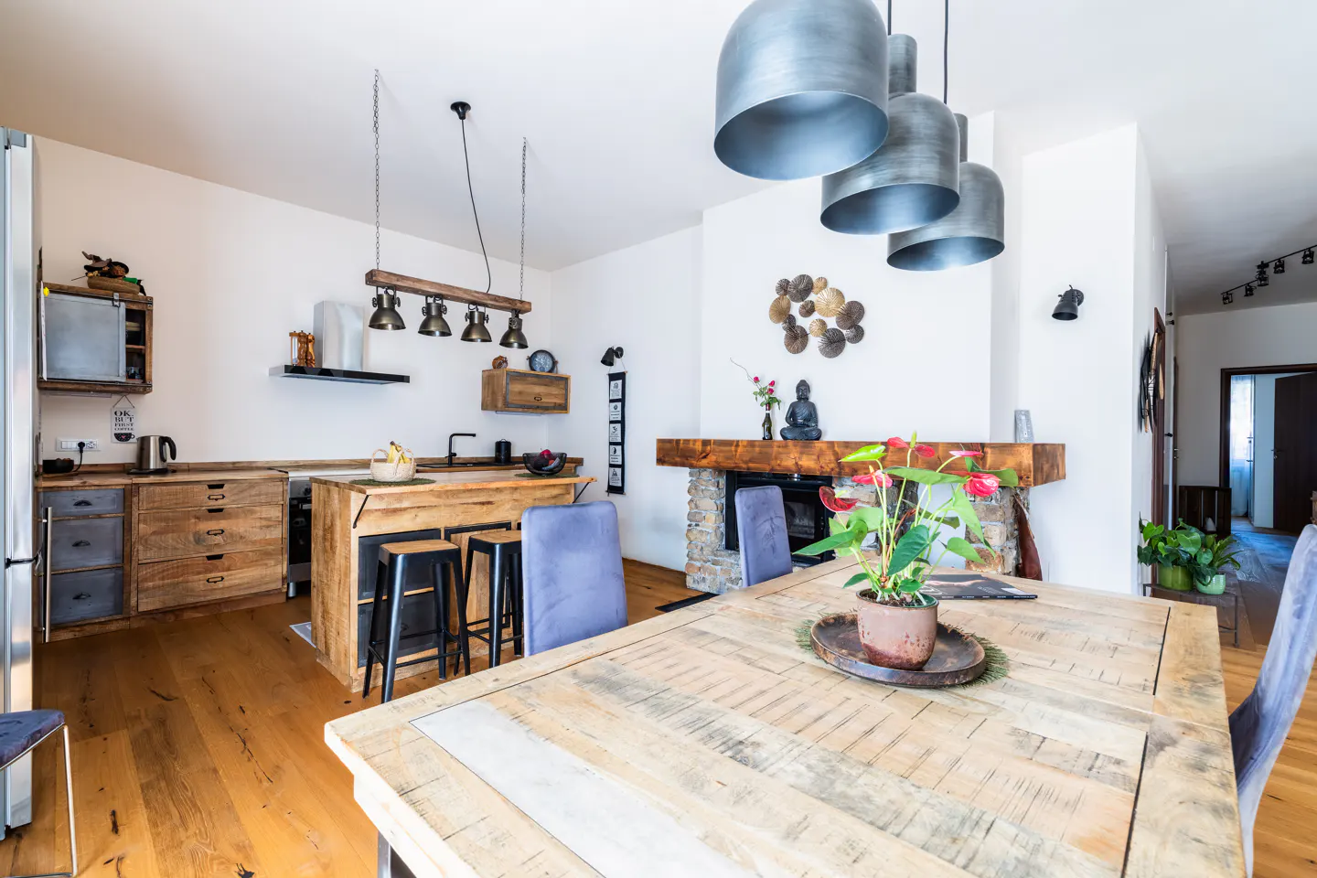 Open-concept kitchen and dining area with rustic wood cabinets, island, table, and chairs. A stone fireplace and metal pendant lights add character.