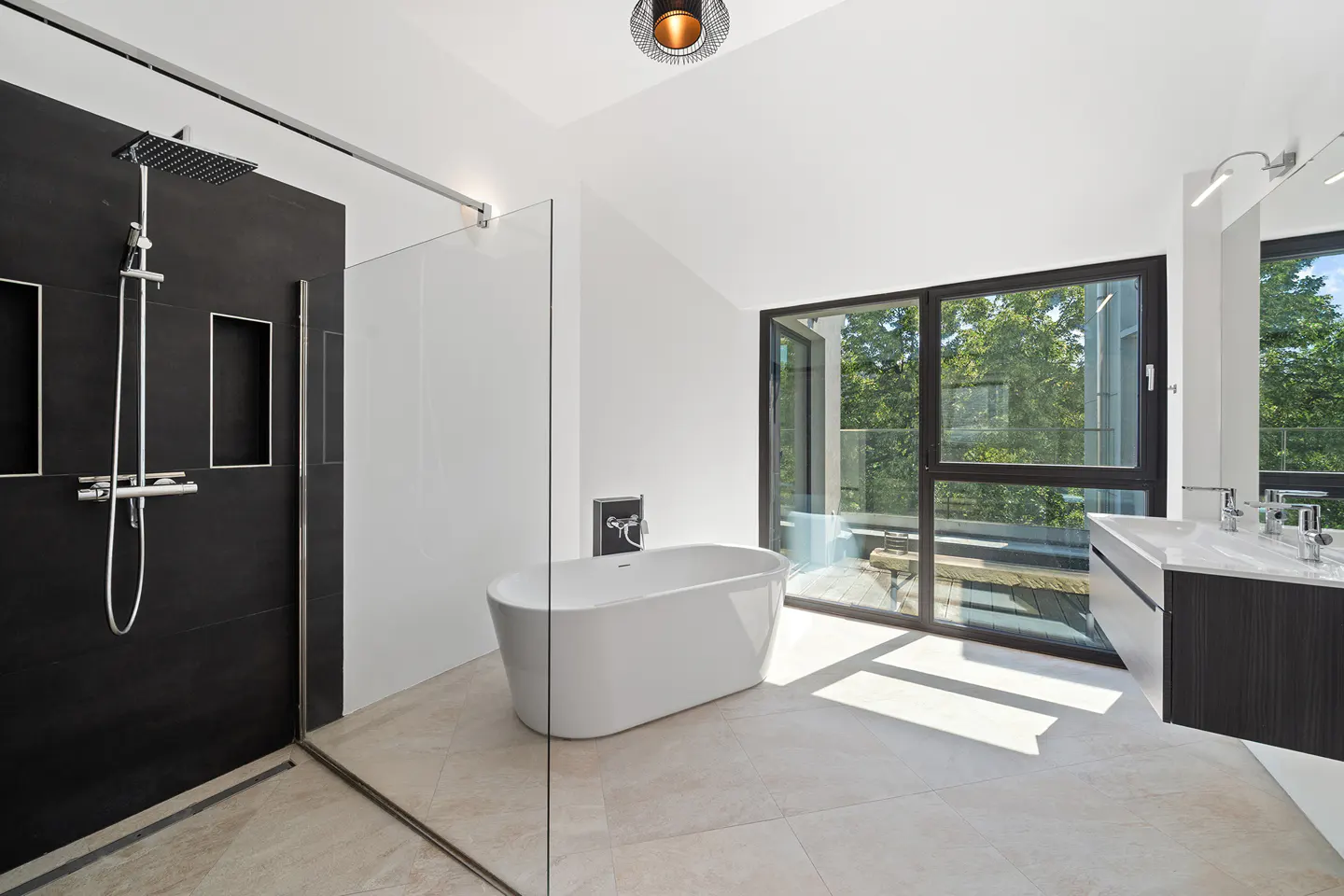 Bright bathroom with a black tiled shower, glass door, white tub, and a large window with trees outside.