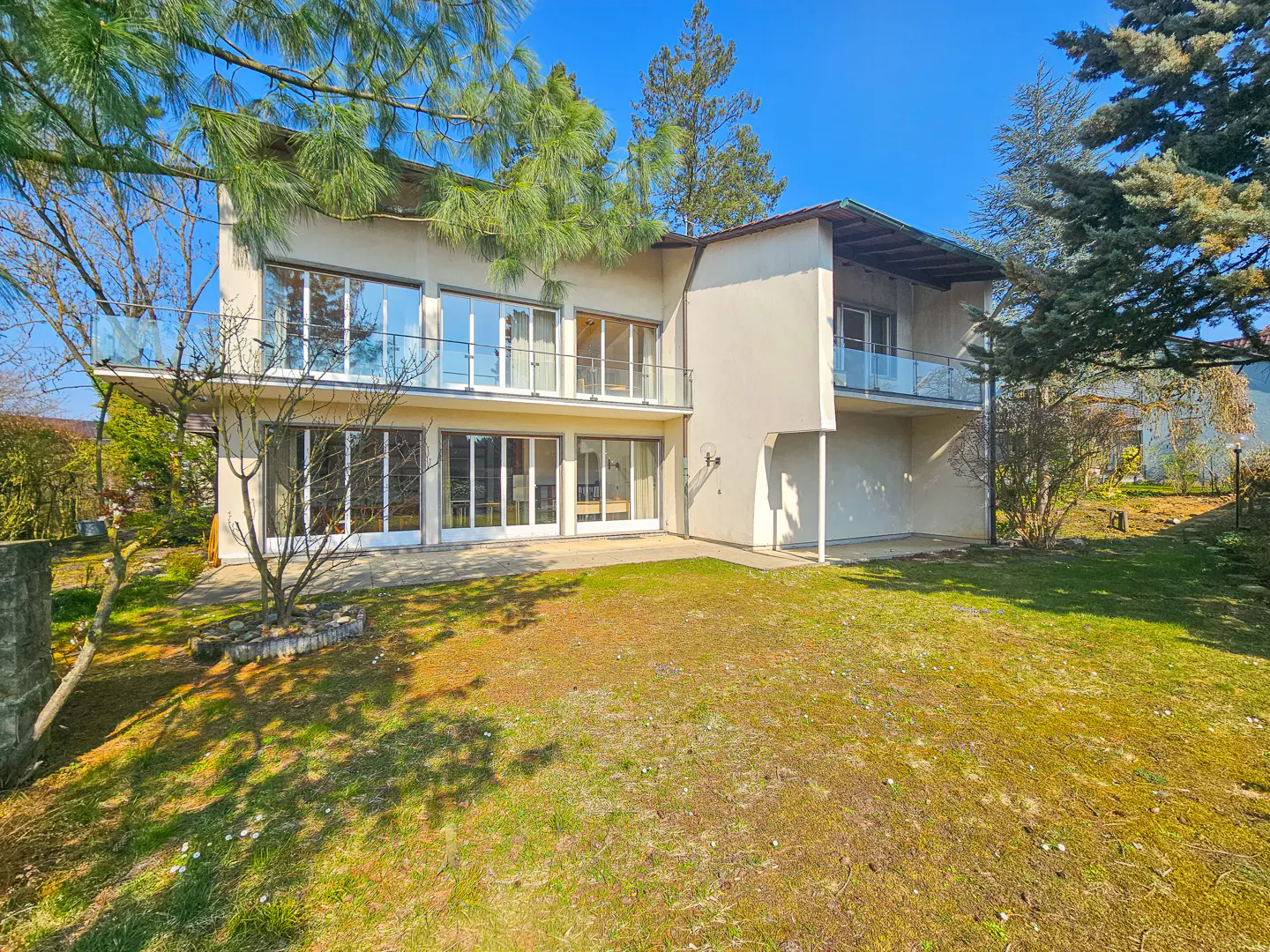 Two-story beige house with large windows and glass balconies, surrounded by green lawn and trees under a blue sky.