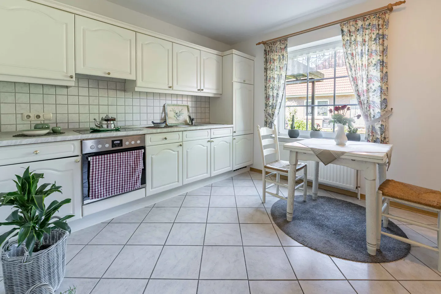 Bright kitchen with white cabinets, tile floor, and a small table with chairs near a window with floral curtains.