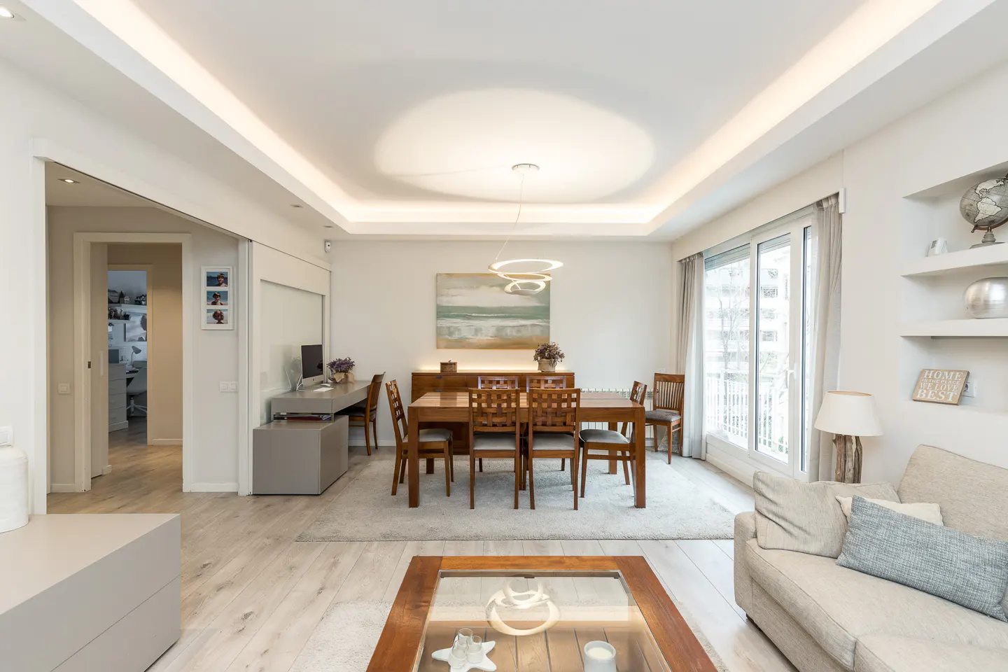 Bright, modern living and dining area with wood table, gray sofa, and light wood floors. A desk and shelves add functional decor.