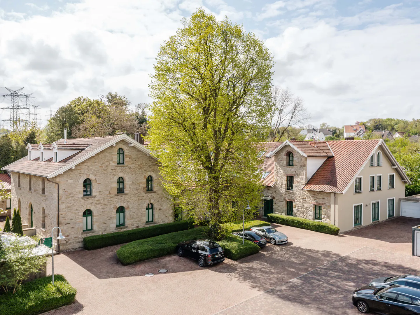 Two stone buildings with red tile roofs flank a large green tree. Cars are parked in the brick courtyard.