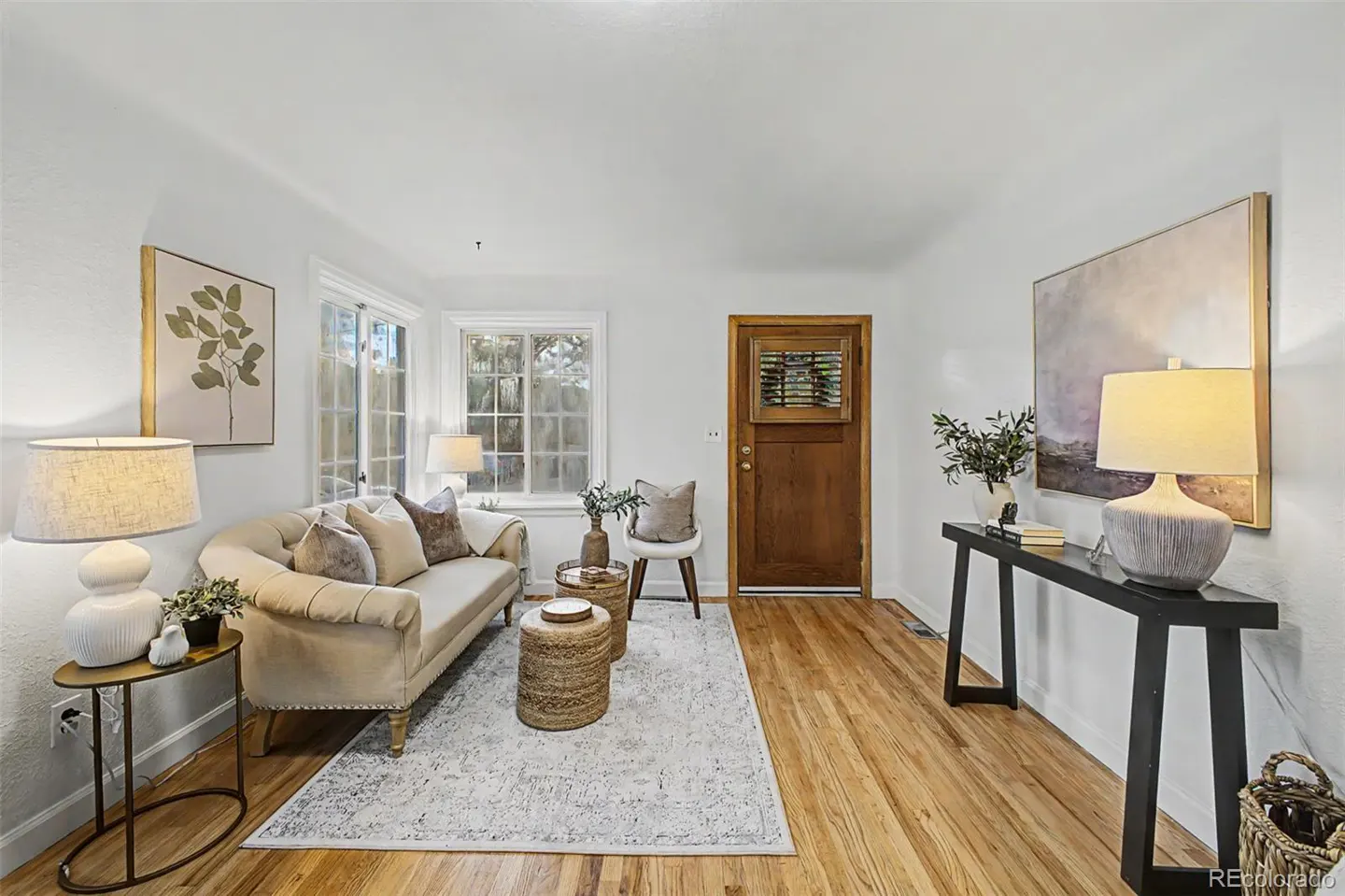 Living room with hardwood floors, white walls, and a beige sofa with throw pillows. A black console table with a lamp sits against the wall.