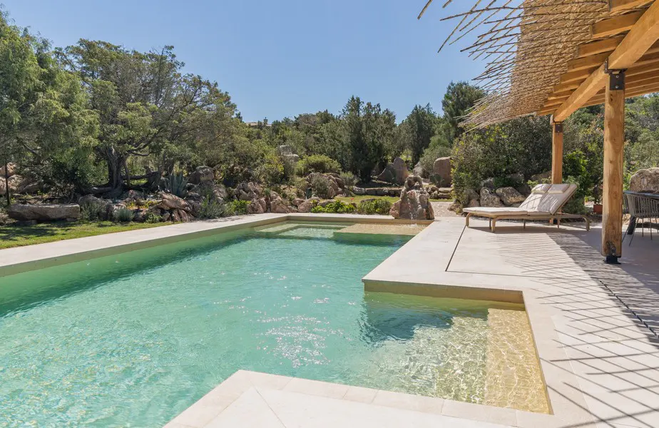 A turquoise pool with a beige deck and lounge chair sits near a wood pergola. Trees and rocks are in the background.