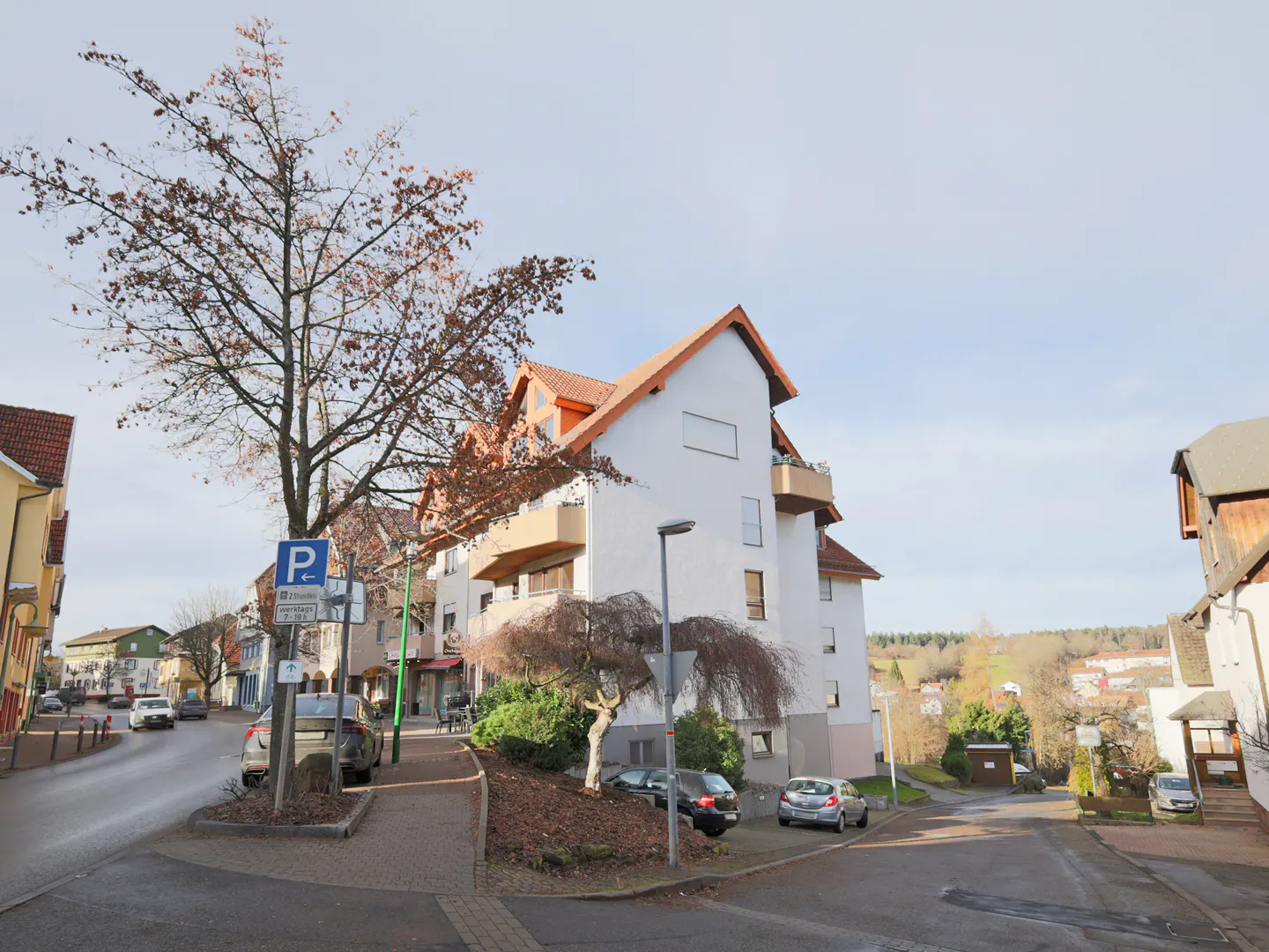 Street view of a white apartment building with a red roof, cars parked on the side of the road, and a tree with brown leaves.