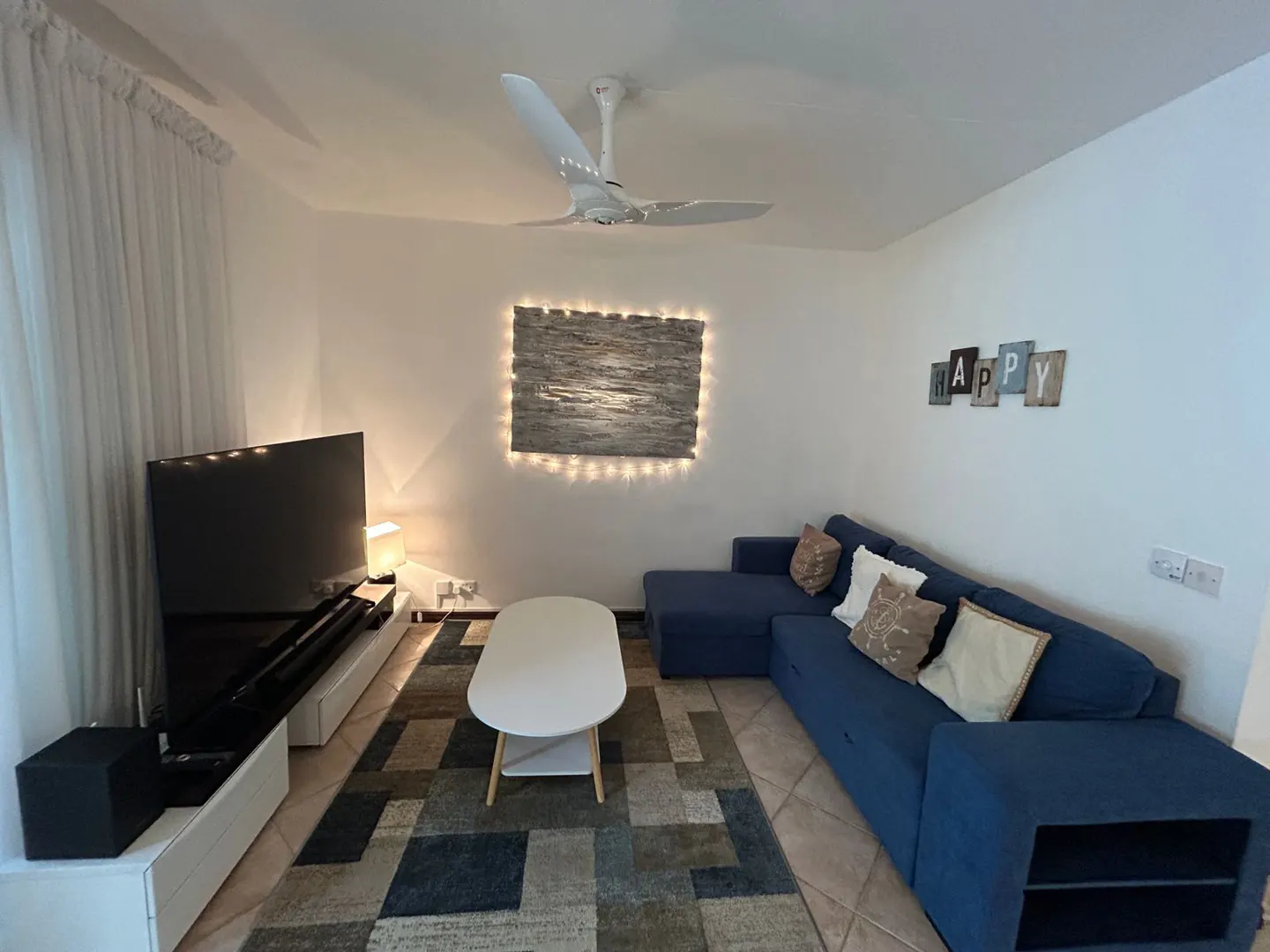 Living room with blue sectional sofa, white coffee table, TV, and a decorative wall art with lights. "HAPPY" sign on the wall.