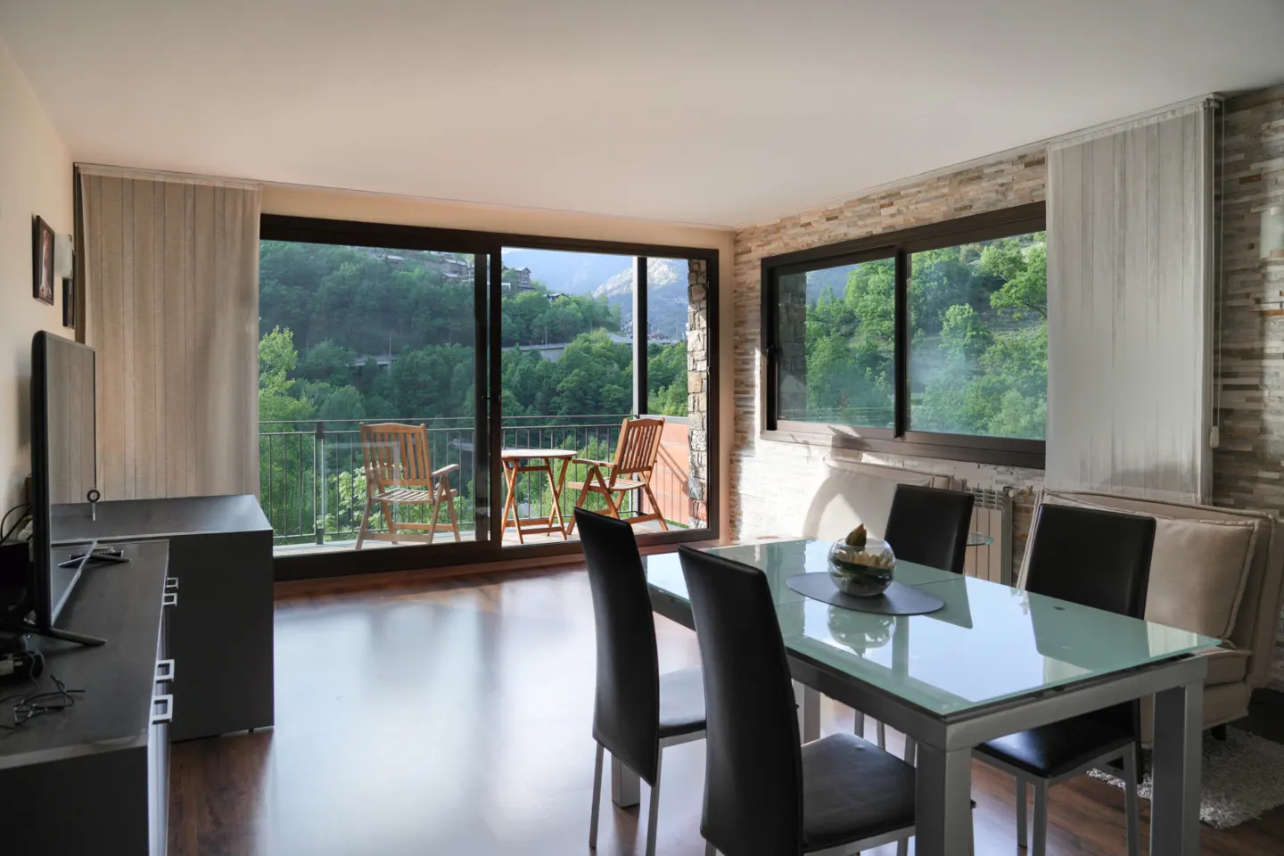 A bright dining room with a glass table and black chairs, a balcony with a view of green hills.