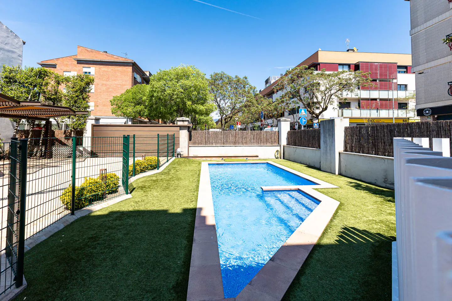 Outdoor pool with blue water and stone trim, surrounded by green artificial turf and fences, with buildings in the background.