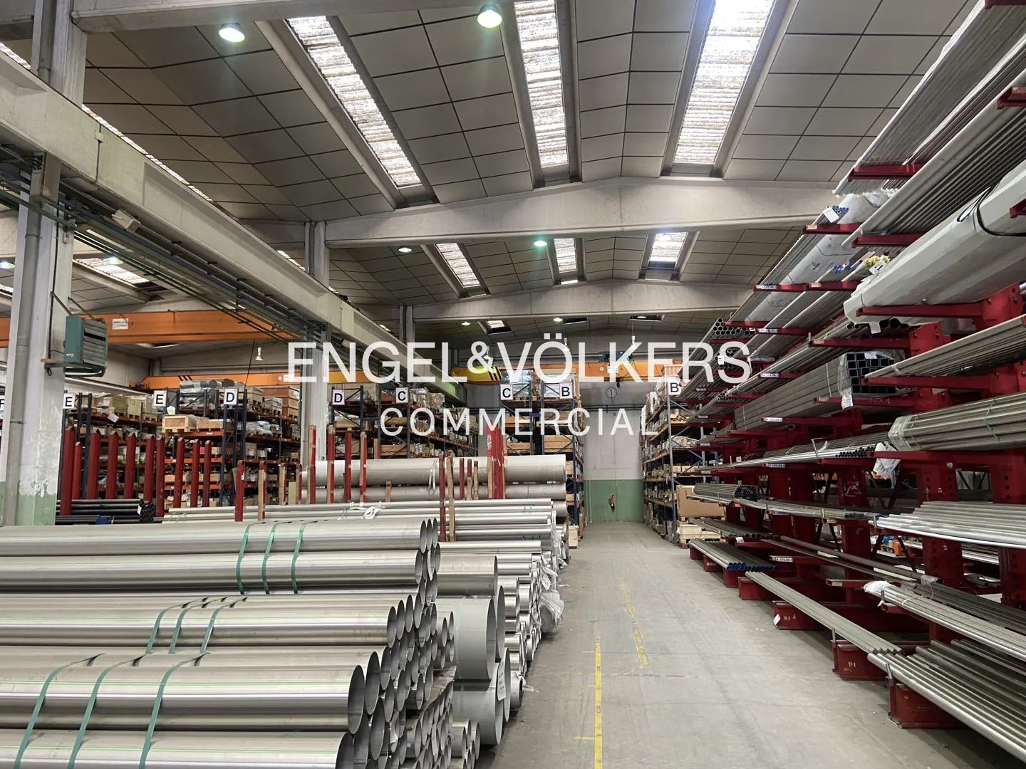 A warehouse interior with metal pipes stacked on shelves. The Engel & Volkers Commercial logo is superimposed in the center.