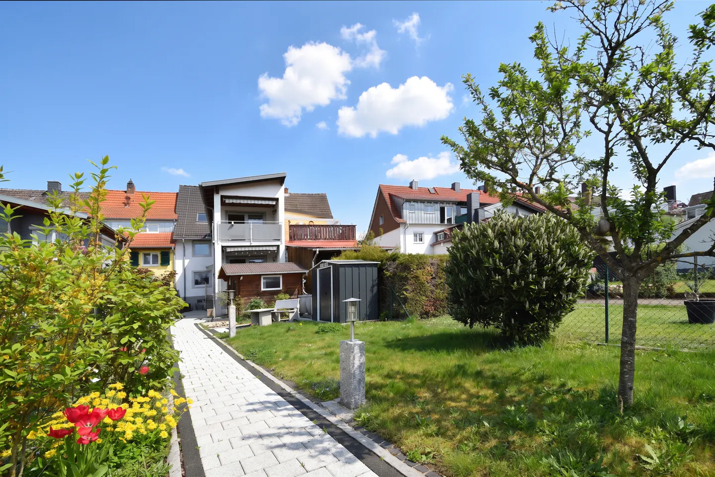 A backyard with a stone path leading to a house with a balcony, green grass, and a blue sky.