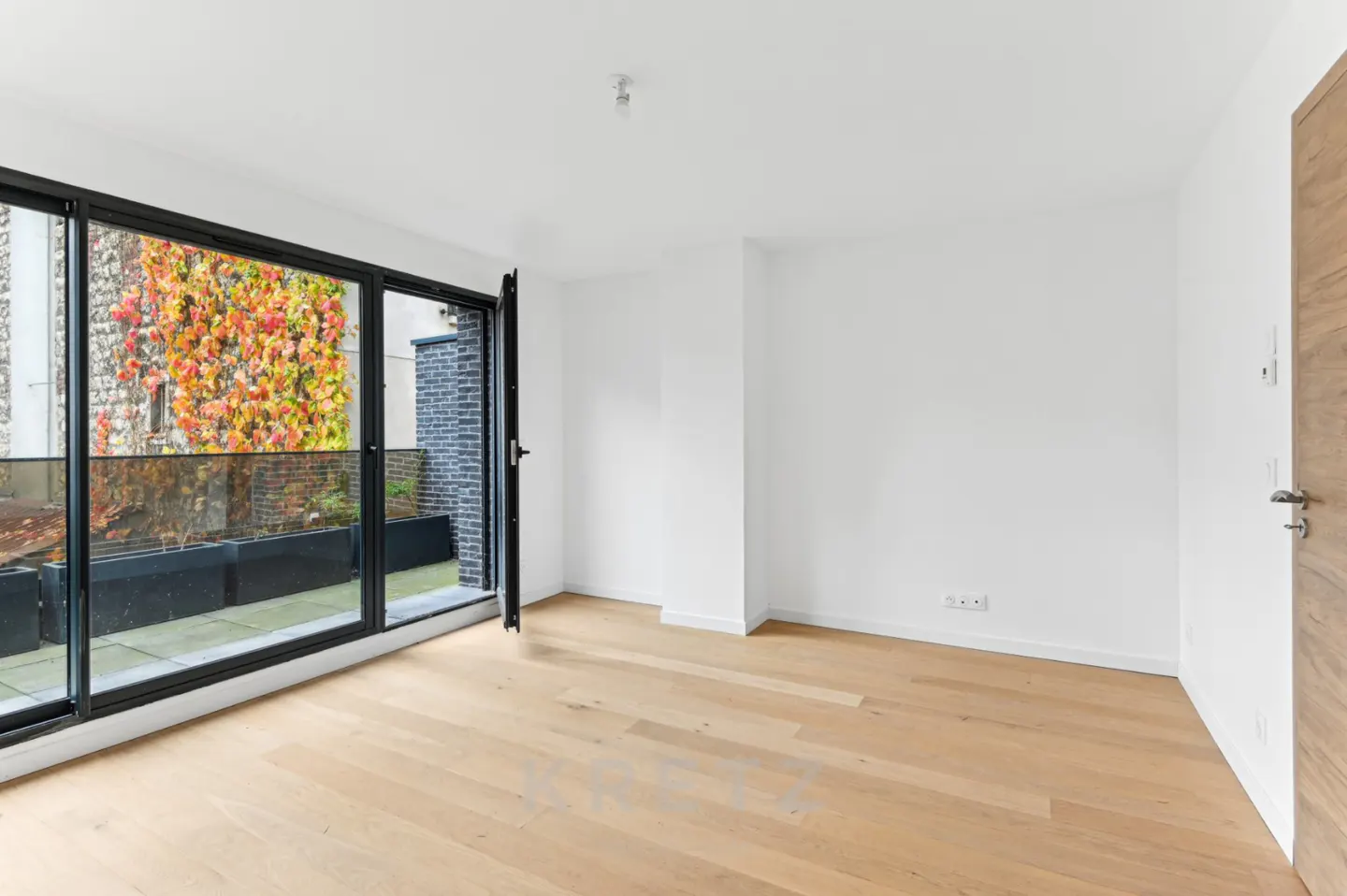 Empty room with light wood floors, white walls, and large glass doors opening to a balcony with autumn-colored vines.