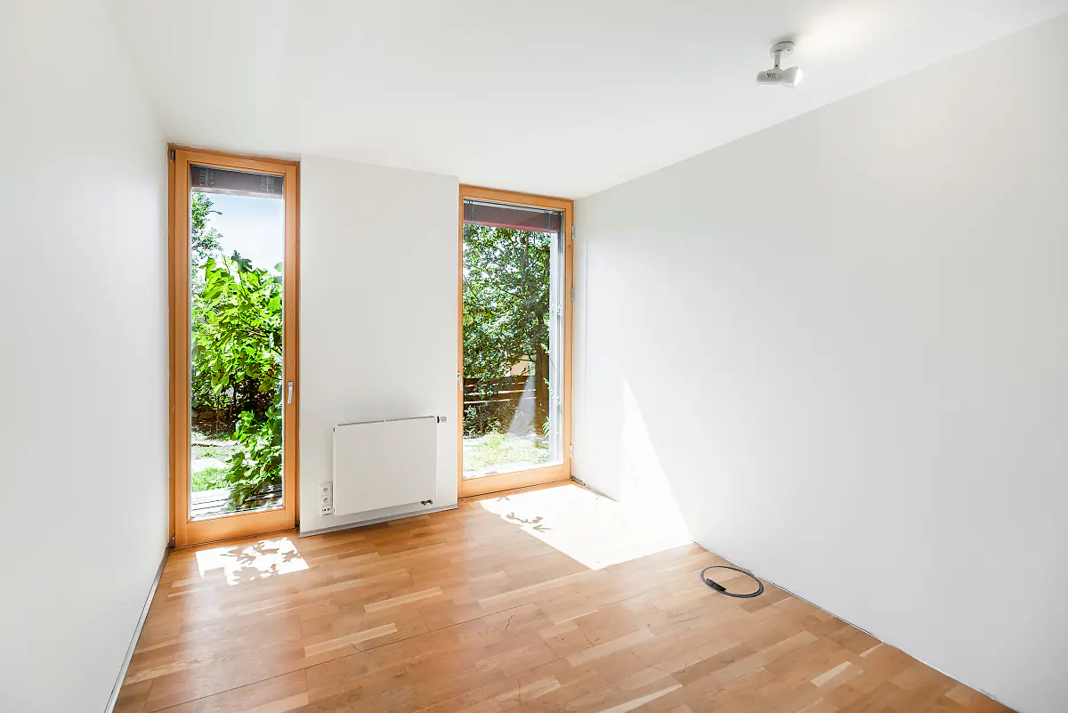 Empty room with white walls, light wood floors, two tall windows with wooden frames, and a white radiator.