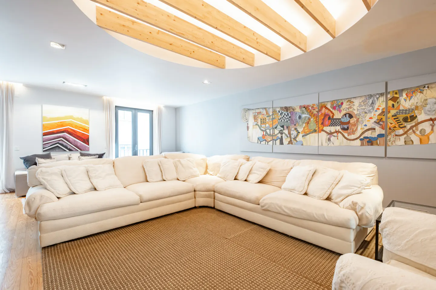 Bright living room with a large white sectional sofa, tan rug, and colorful art. The ceiling has wooden beams and a circular opening.