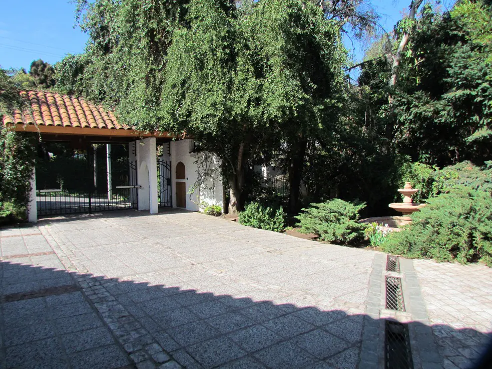 Gated driveway with a red tile roof over the entrance. Lush green trees and bushes surround the property. A small fountain is visible.