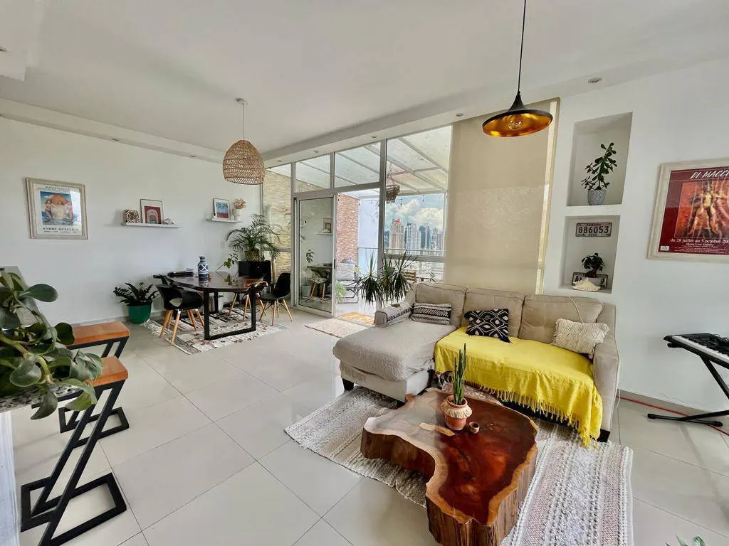 Bright, airy living space with white tile floors, a sectional sofa with a yellow throw, and a unique wood coffee table. Dining area and balcony visible.