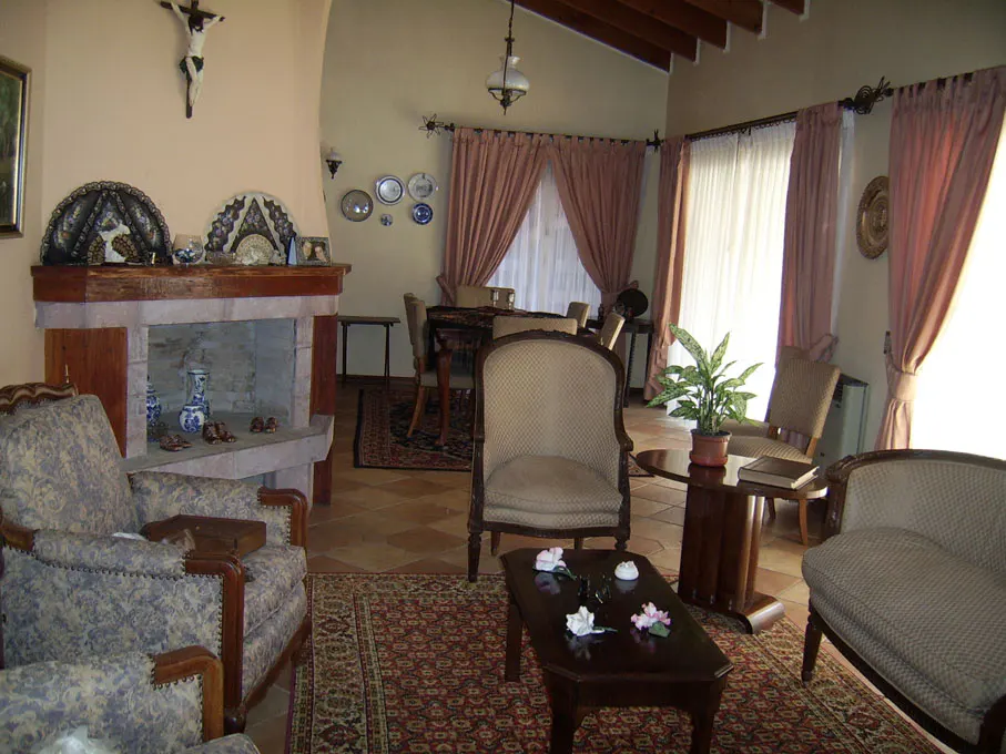 A living room with armchairs, a fireplace, and pink curtains. A crucifix hangs above the fireplace.
