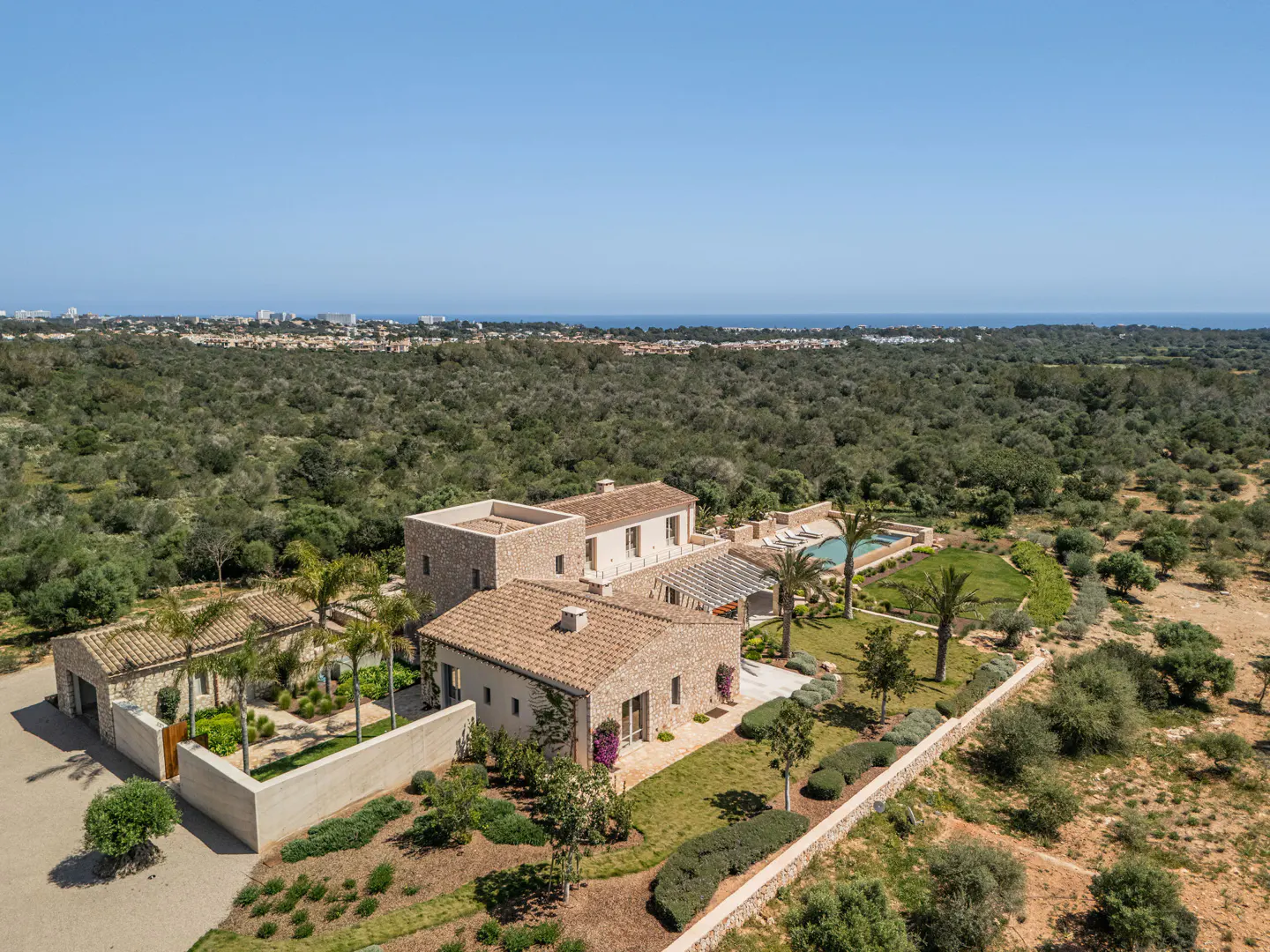 Aerial view of a stone villa with a tiled roof, pool, and palm trees, surrounded by a vast olive grove near the coast.