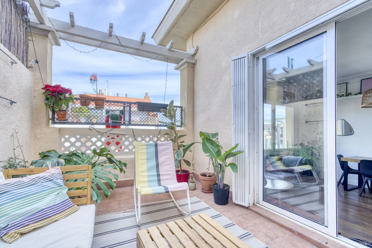 A sunlit balcony with plants, a striped chair, and a wooden pallet table. Sliding glass doors lead to an interior dining area.
