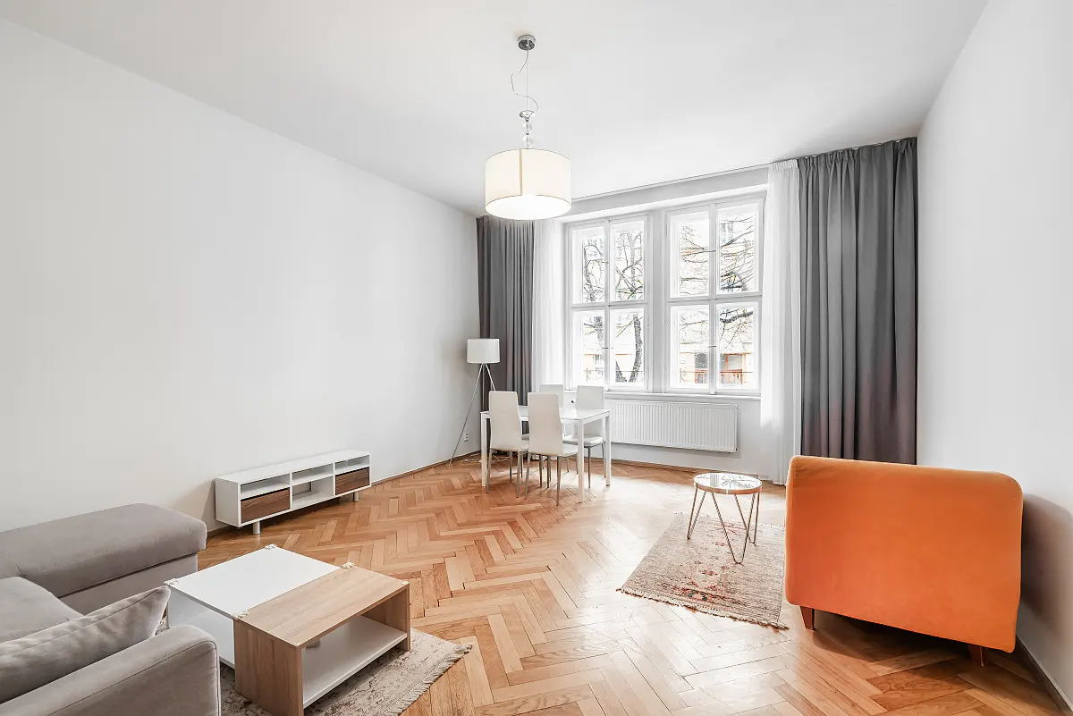 Bright living room with herringbone wood floors, white walls, and gray curtains. A white table and chairs sit near a large window.