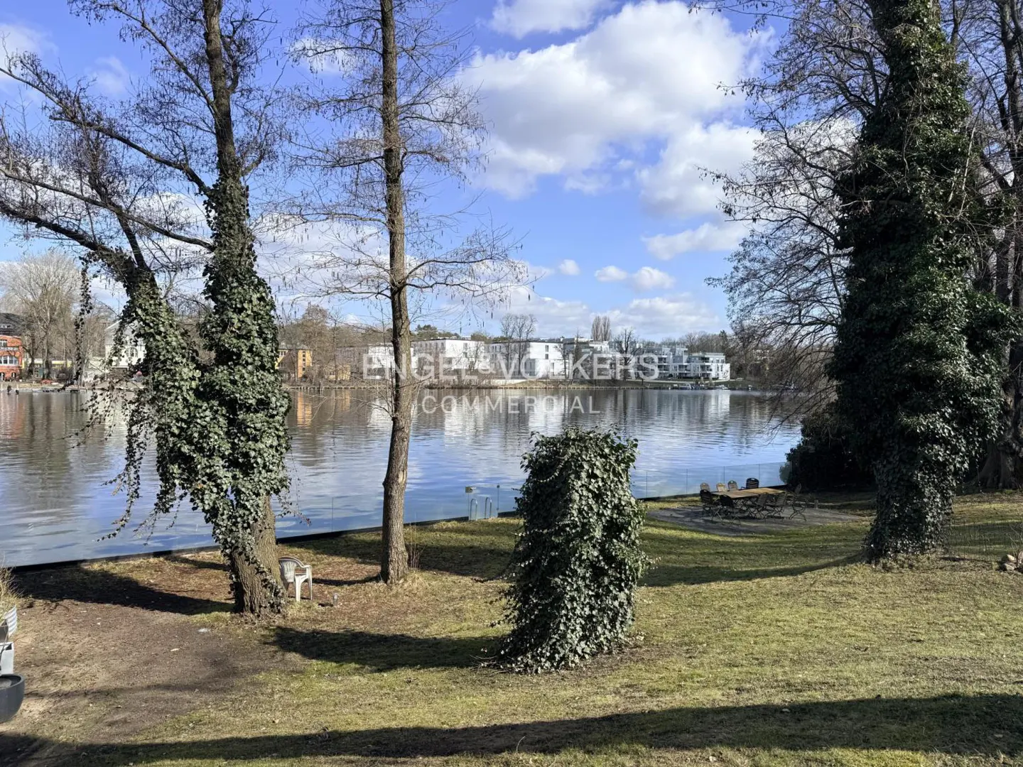 Lakeside view with trees, green grass, and a glimpse of buildings across the water under a partly cloudy sky.